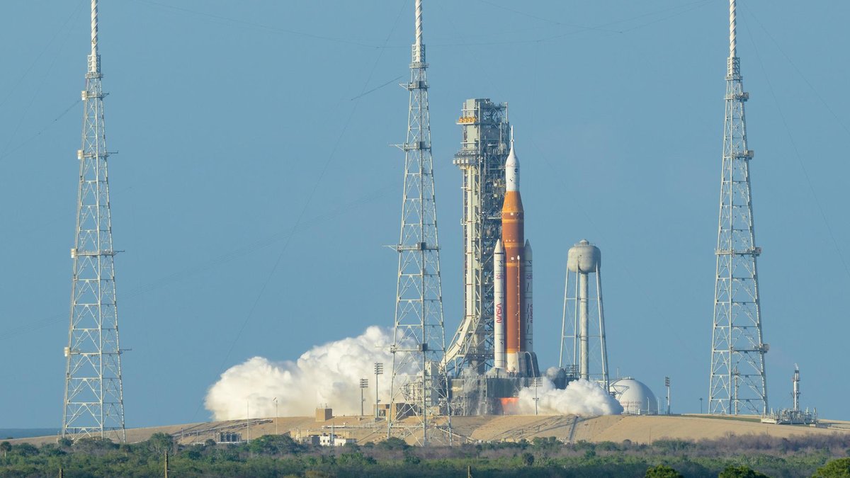 Orange and white SLS rocket lifting off from a launch pad between lightning towers, with clouds of exhaust at its base