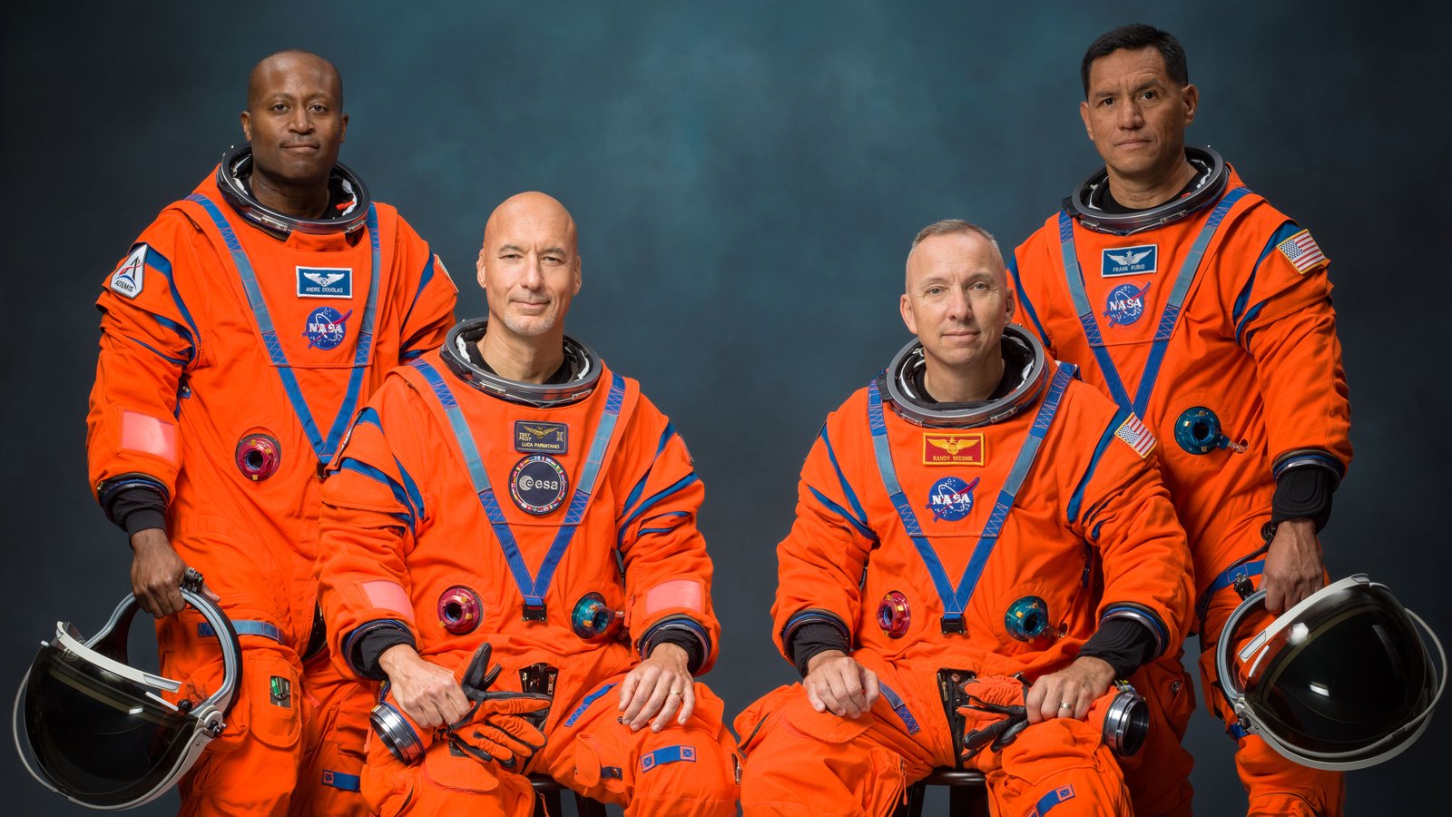 The four Artemis III astronauts in orange flight suits in a studio portrait. Andre Douglas and Frank Rubio stand at the back holding their helmets, with Luca Parmitano and Randy Bresnik seated in front of them