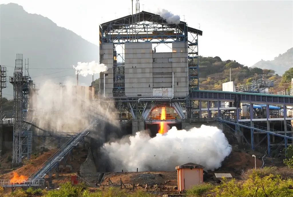 A cryogenic rocket stage firing inside a tall test stand on a hillside, with a bright flame at its base and clouds of white vapour