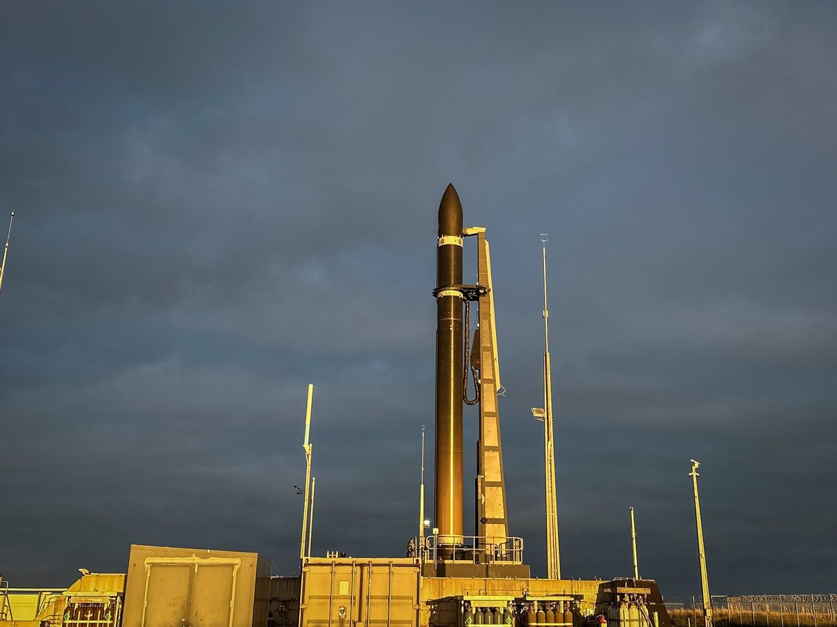 An Electron rocket standing upright on a launch pad, lit gold by low sun under a dark, cloudy sky
