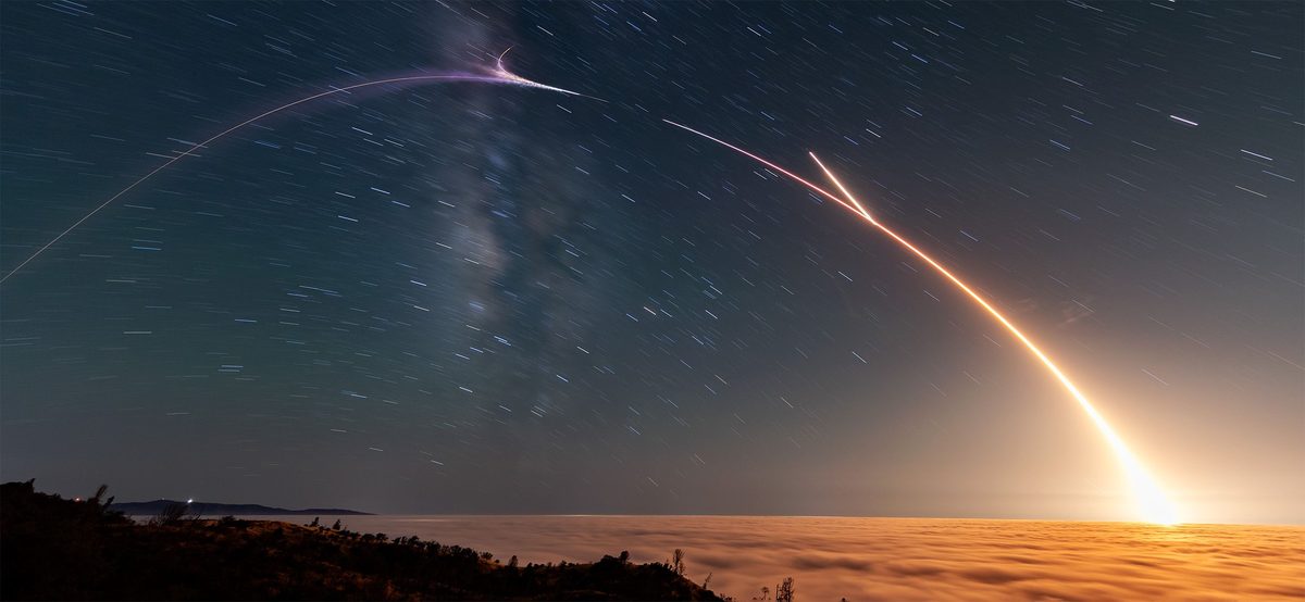A long-exposure night photo of a Falcon 9 launch, with a bright curved streak of light rising from a sea of glowing cloud under a starry sky and the Milky Way