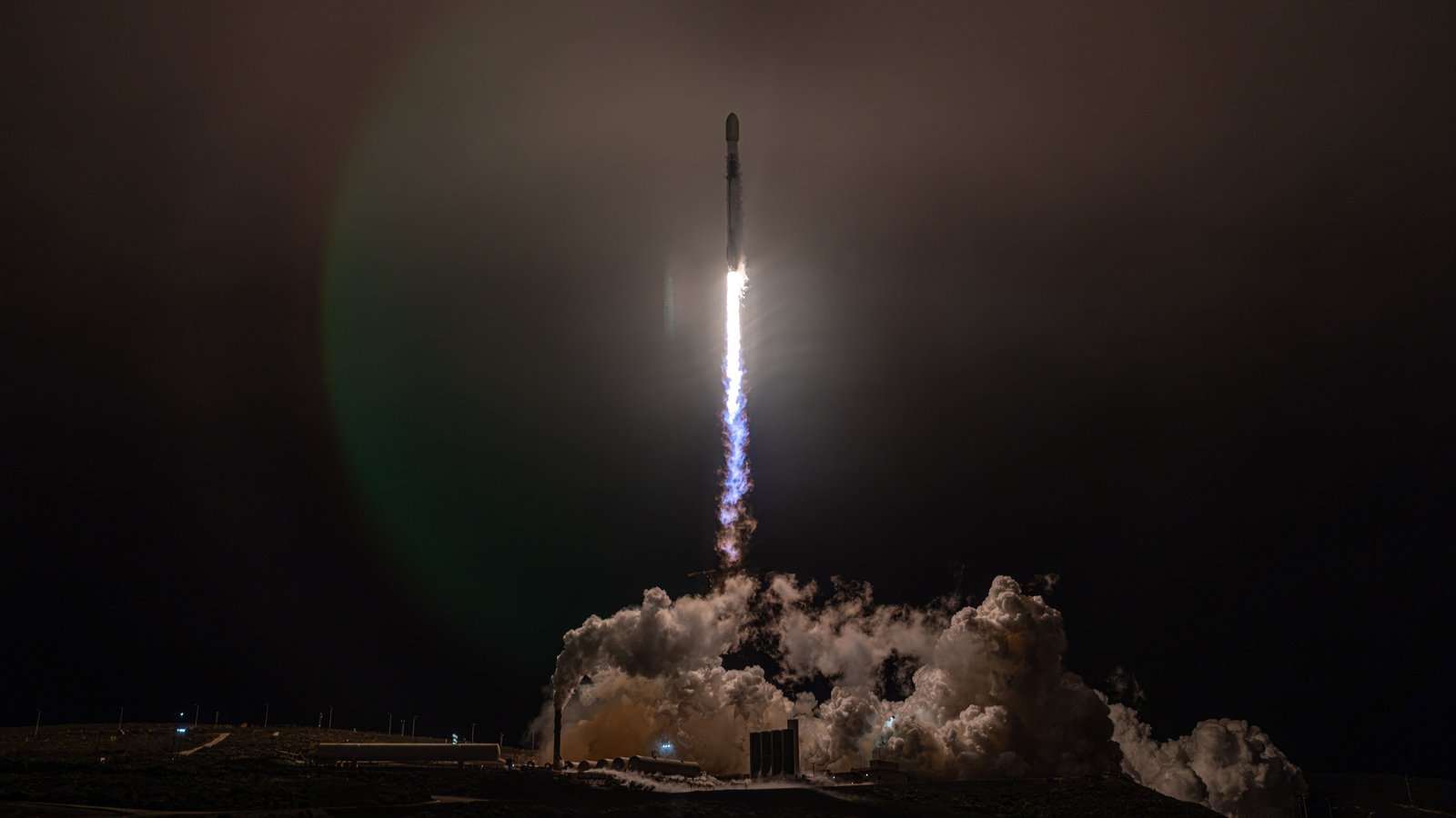 A Falcon 9 rocket climbing on a bright flame into the night sky above Vandenberg, lighting the clouds of exhaust below it