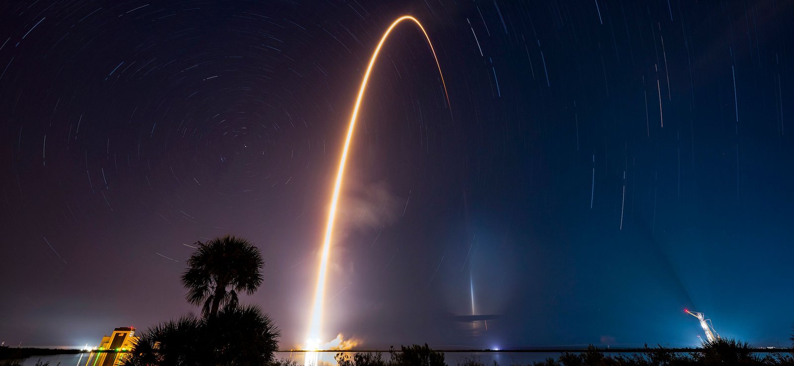 A long-exposure photo of a Falcon 9 launch, a bright curved streak rising into the night sky
