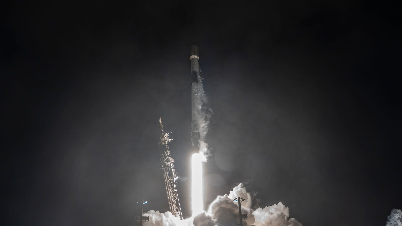 A Falcon 9 rocket lifting off at night on a bright flame, lighting up the launch pad and smoke
