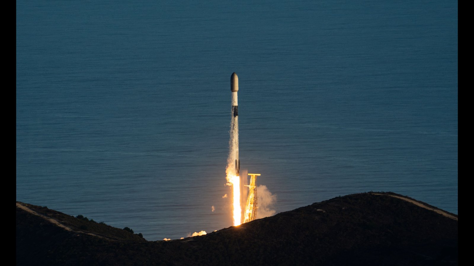A Falcon 9 rocket rising from its launch pad on a bright flame, with the Pacific Ocean behind it