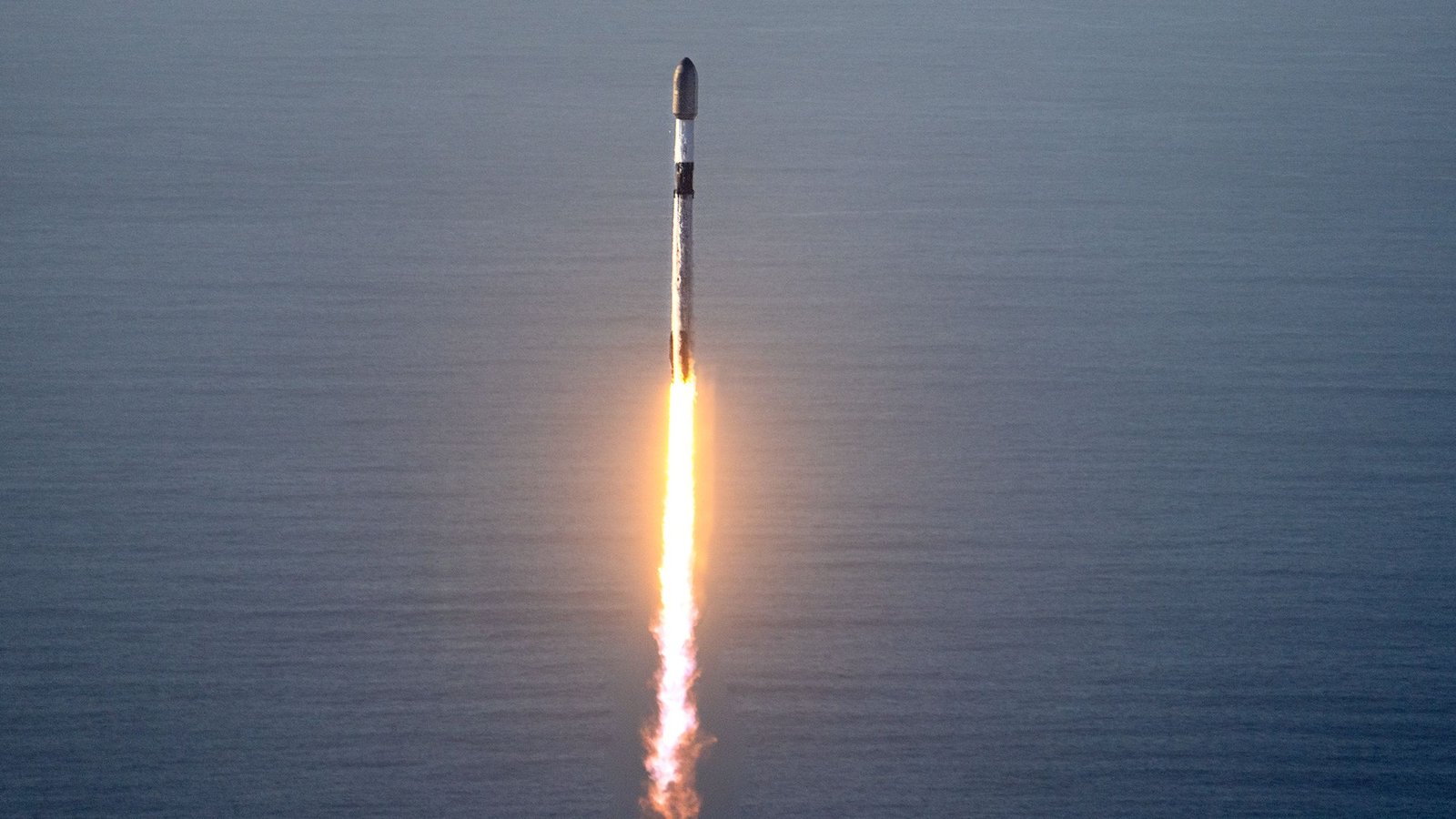 A Falcon 9 rocket climbing on a bright flame above the coast, with the ocean in the background