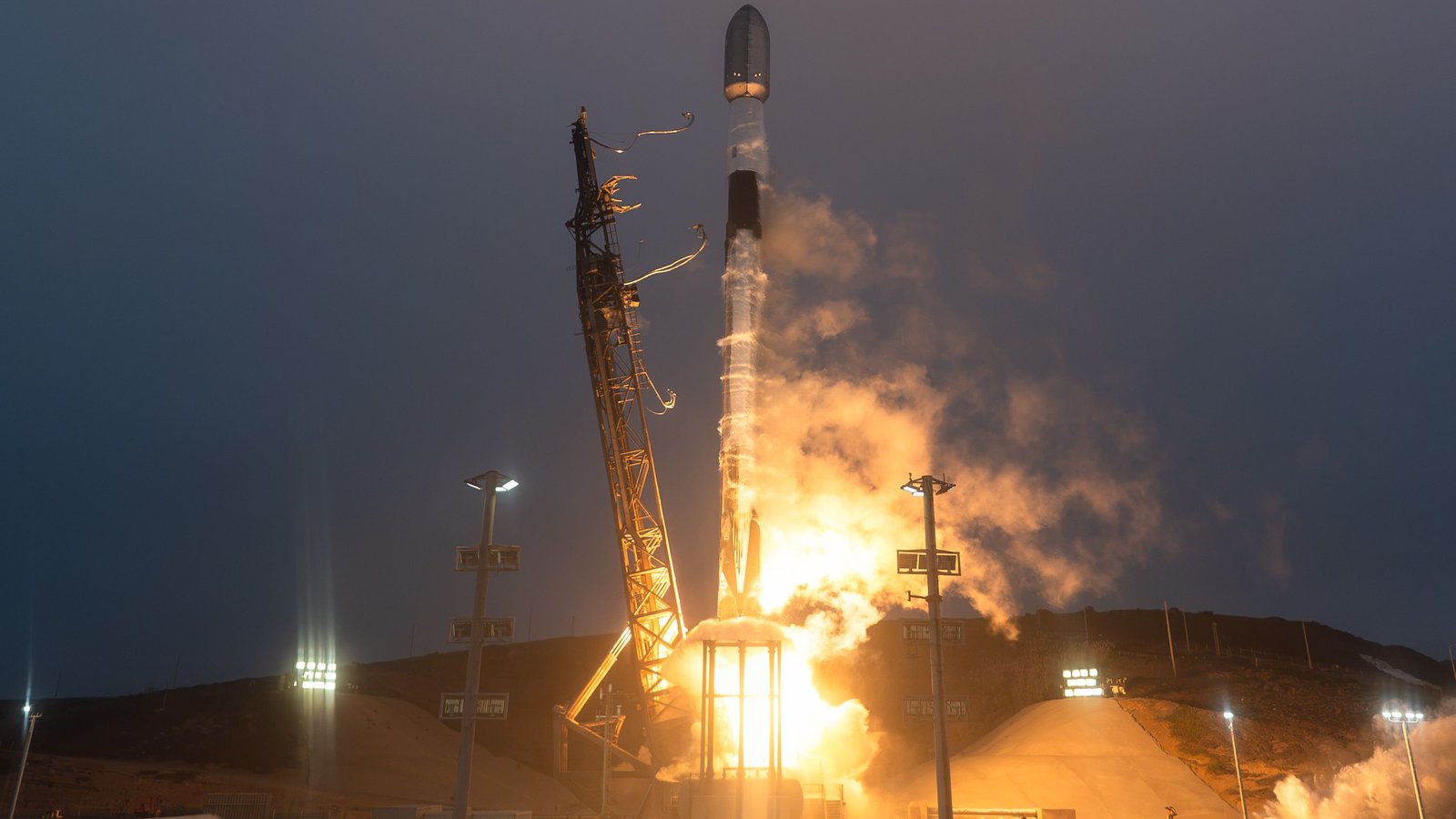 A Falcon 9 rocket rising on a bright orange flame beside its launch tower at dusk, with clouds of exhaust lit orange around the pad