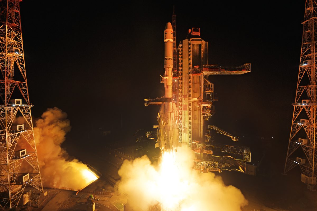 A GSLV rocket lifting off in the dark before dawn, lit by its own exhaust, beside the launch tower at Sriharikota