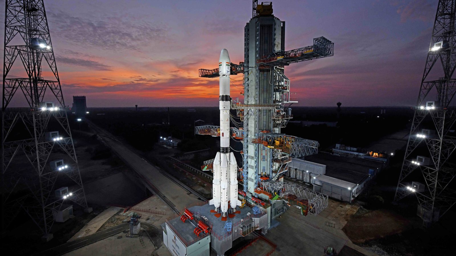 ISRO's GSLV-F18 rocket standing beside its service tower on the Second Launch Pad at Sriharikota under an orange and purple dusk sky, with floodlight towers on both sides
