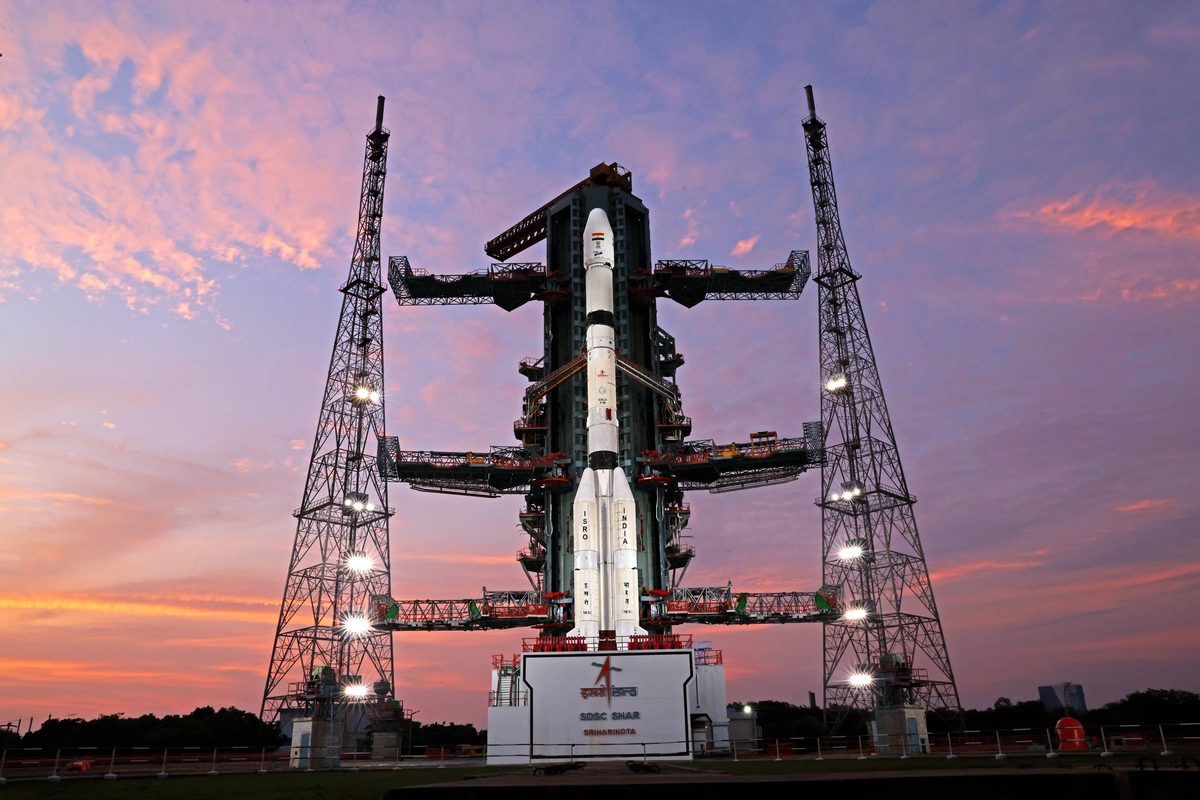GSLV-F18 seen head-on between two tall lattice lightning towers, with its service tower arms open, against a pink and orange sunset sky