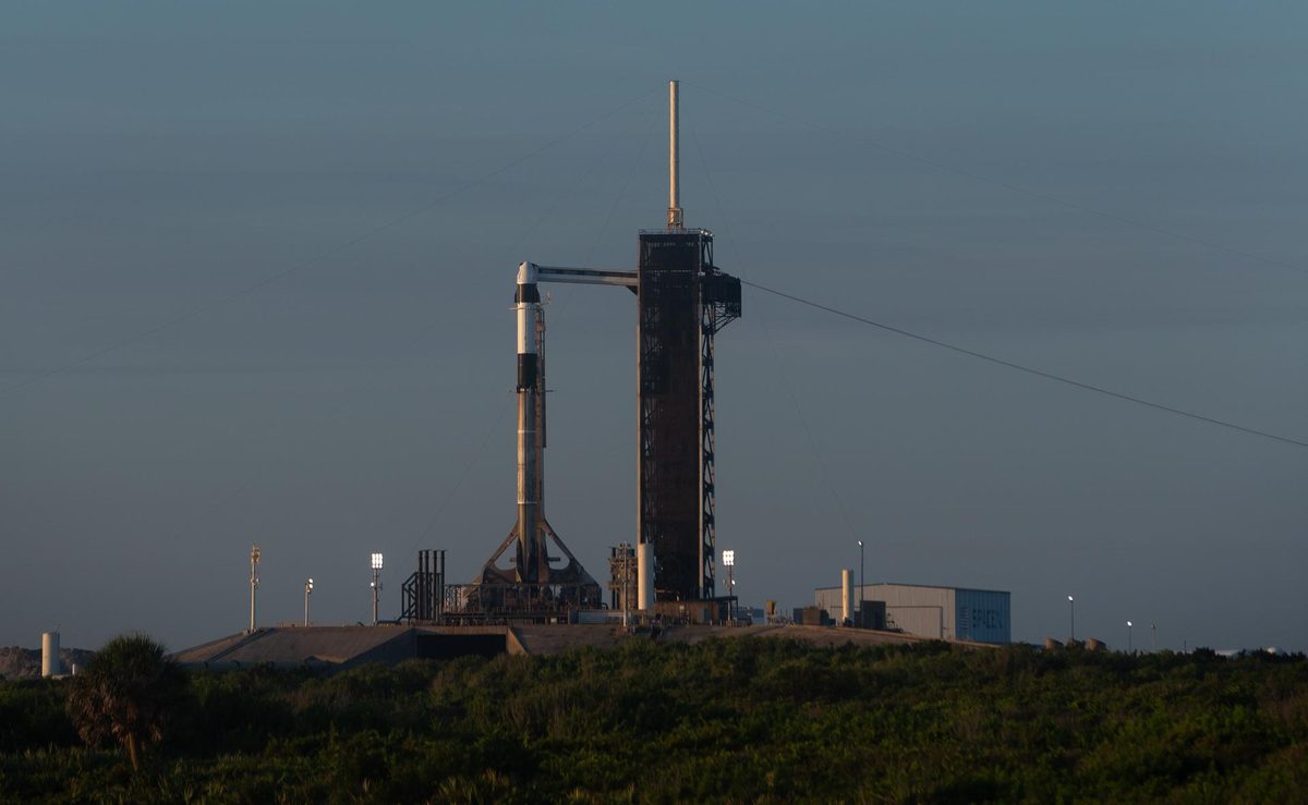 A Falcon 9 rocket with a Dragon capsule on top standing beside the tall launch tower at Launch Complex 39A in the morning light, with scrubland in front