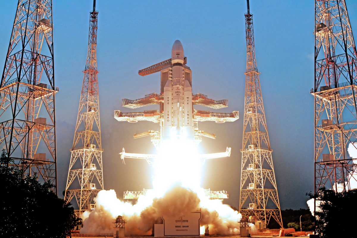 An LVM3 rocket lifting off from Sriharikota, its two large side boosters firing, between tall lightning towers against a blue evening sky