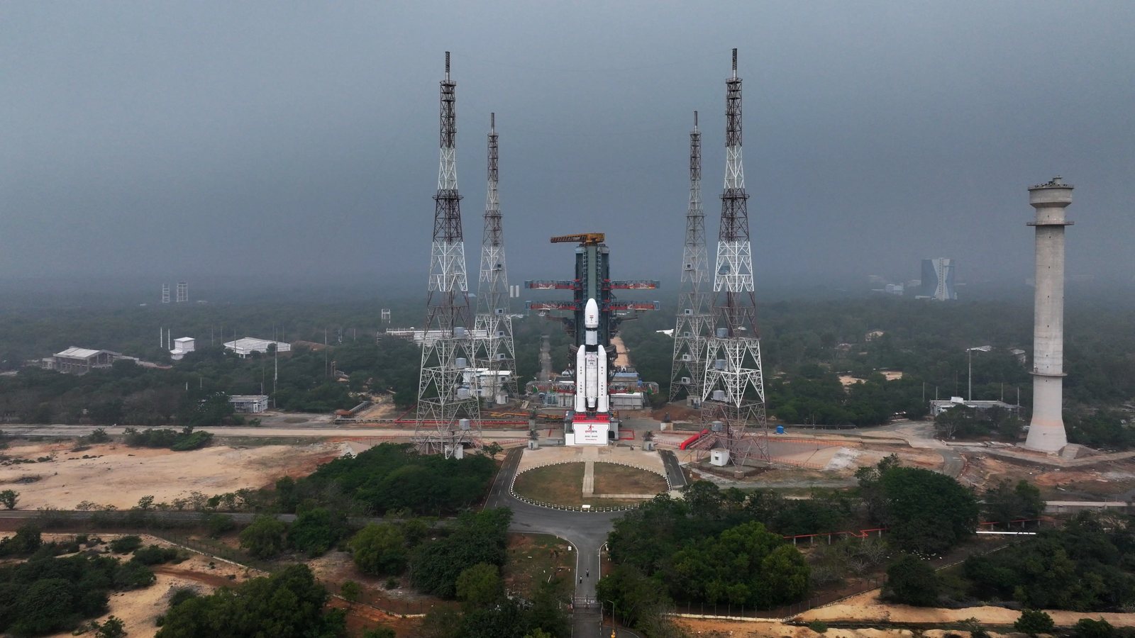 Aerial view of a white LVM3 rocket standing at its launch pad at Sriharikota, between four tall steel towers, with forest around it and a grey sky