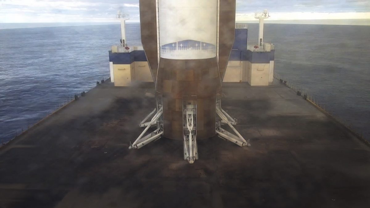 The base of a New Glenn booster standing on its landing legs on the dark deck of Blue Origin's landing ship, with open ocean on both sides