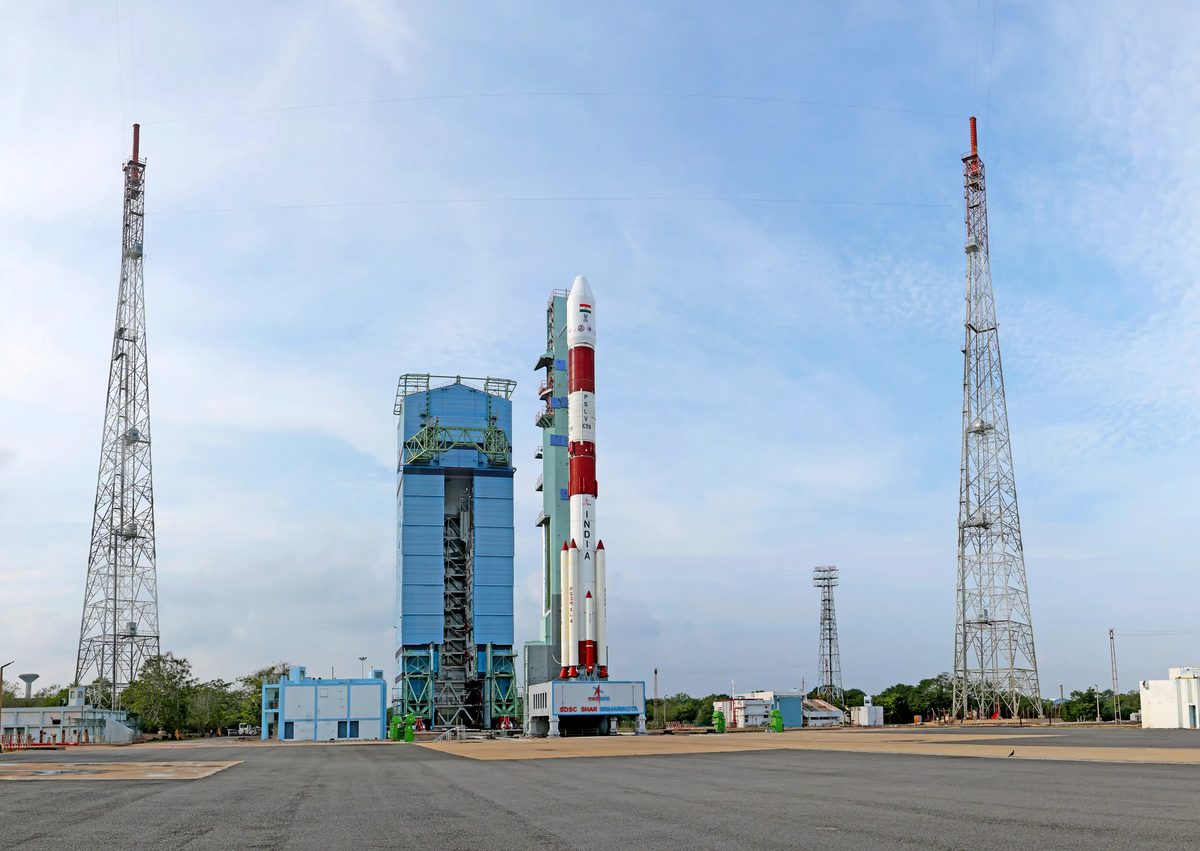 A white and red PSLV rocket standing on its launch pad at Sriharikota beside the tall blue service tower, with lightning towers on either side and a blue sky