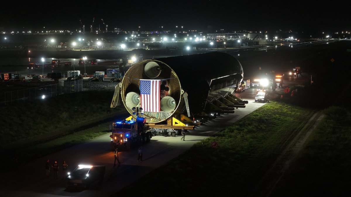 A Starship upper stage lying on a transport vehicle at night, with work lights around it and a US flag painted on its side