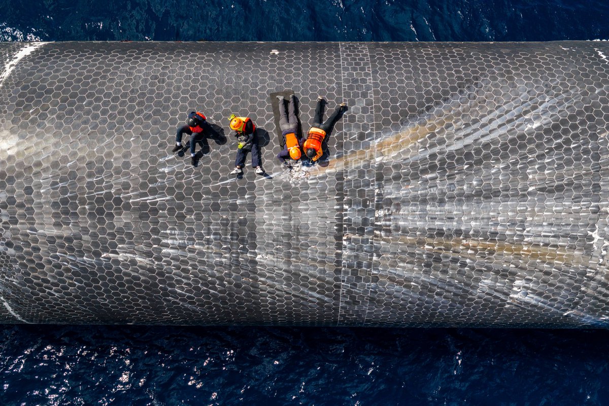 Seen from directly above, four SpaceX crew members in life jackets kneel and lie on the curved black heat-shield tiles of a floating Starship, with dark blue sea all around