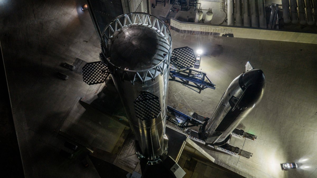 Looking down at night from high on the launch tower: the top of the Super Heavy booster with its grid fins in the foreground, and the ship lying on a transport stand beside it
