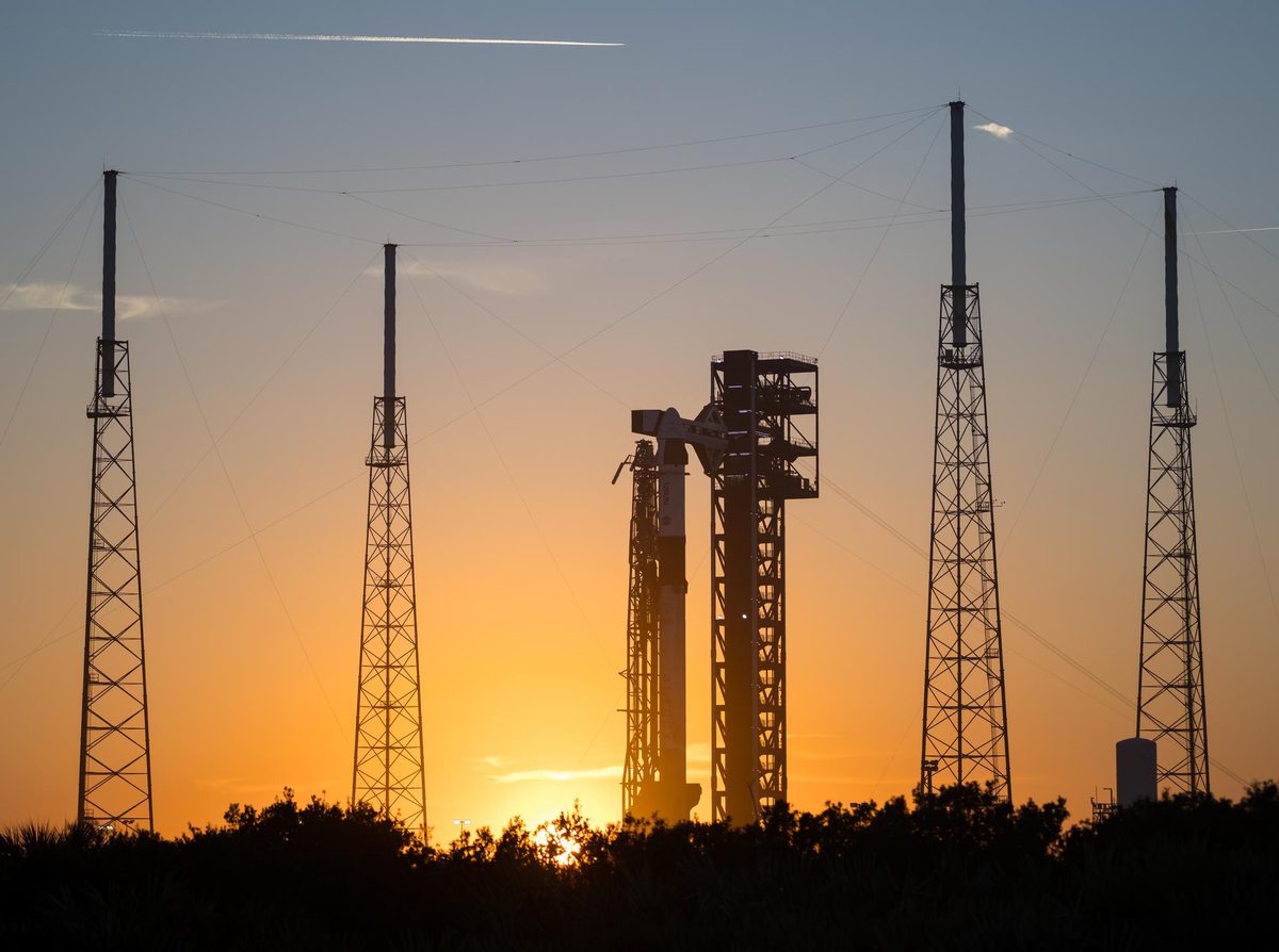 A Falcon 9 rocket with a Dragon capsule on top standing beside its tower at Space Launch Complex 40, silhouetted against an orange sunset sky