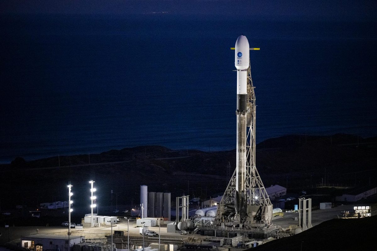 A Falcon 9 rocket standing on a floodlit launch pad at night on the California coast, with the dark Pacific behind