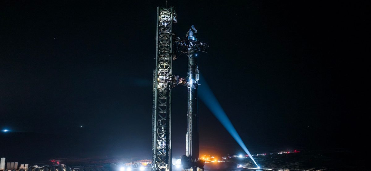 Starship standing beside its launch tower at night at Starbase, lit by floodlights, with a searchlight beam crossing the sky