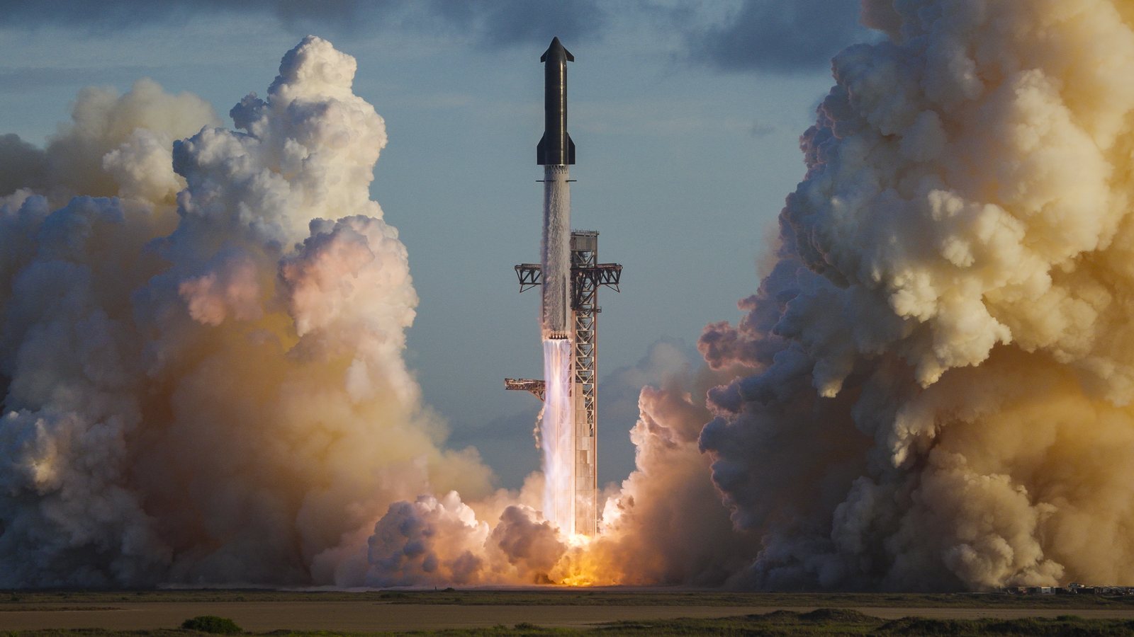 Starship's full stack, the ship on top of the Super Heavy booster, lifting off beside the launch tower at Starbase on Flight 14 between huge clouds of exhaust