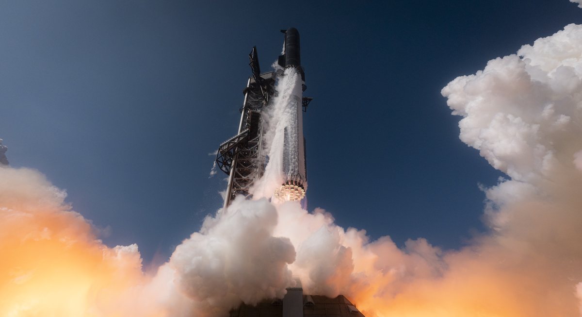 Starship V3 lifting off beside its launch tower, with orange-lit clouds of exhaust boiling out across the pad
