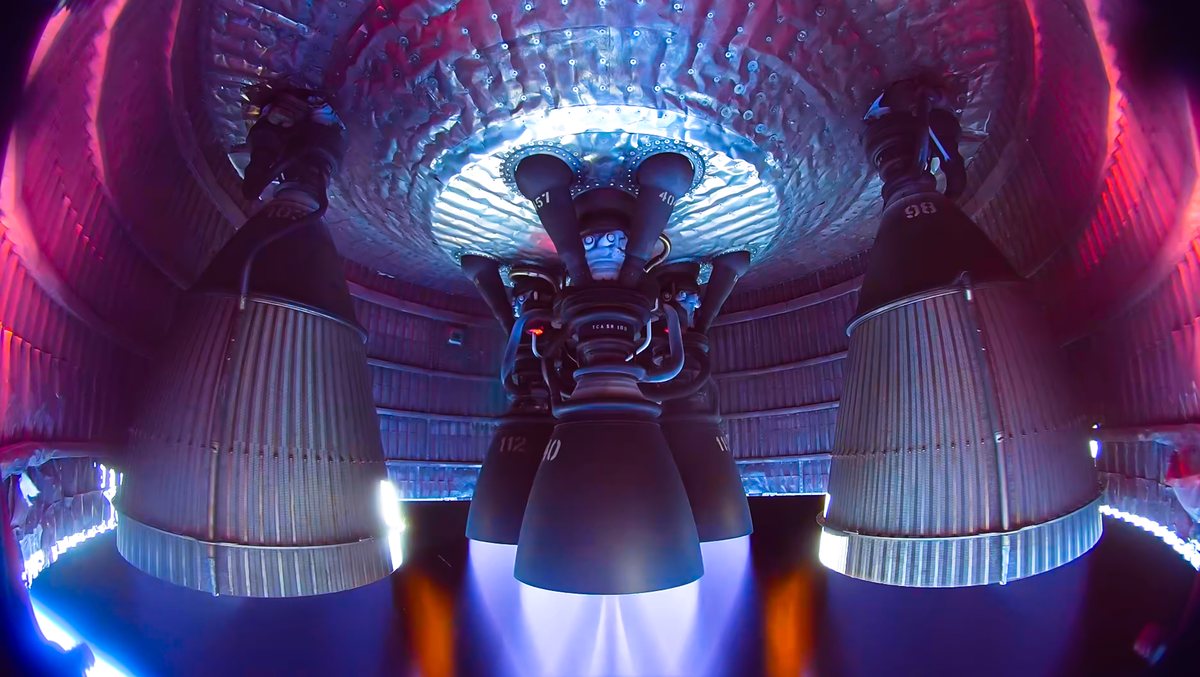 Onboard camera view inside the engine section of a Starship V3 vehicle, looking at Raptor engines firing, their large bells glowing at the rims under pink and blue light