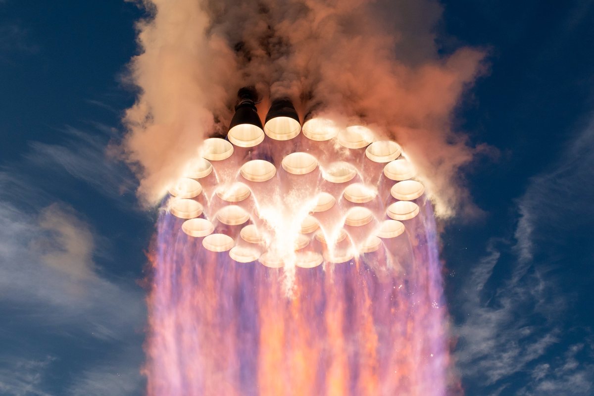 Looking up at the base of the Super Heavy booster as its ring of engines fires against a blue sky