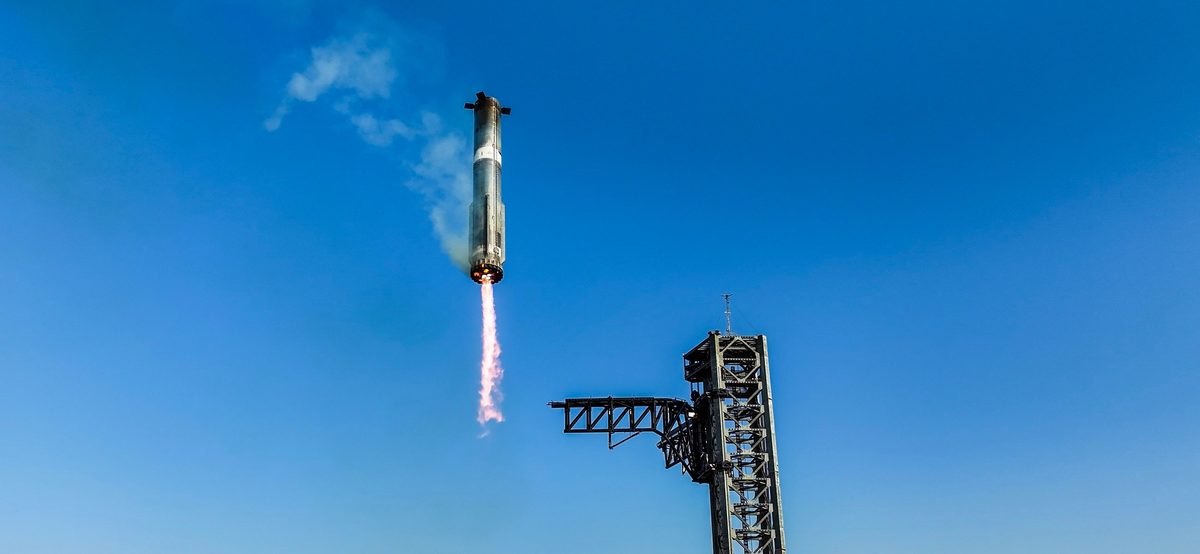 The Super Heavy booster descending tail-first against a blue sky, engines firing, above the launch tower with its arms held out