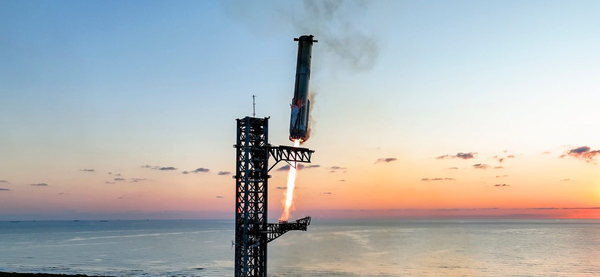At sunrise over the sea, the Super Heavy booster lowers itself beside the launch tower on a column of engine exhaust, between the tower's two arms