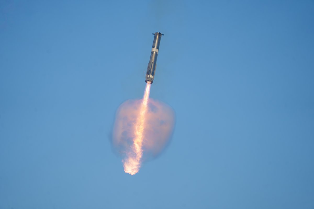The Super Heavy booster descending against a blue sky with a bright plume of engine exhaust beneath it