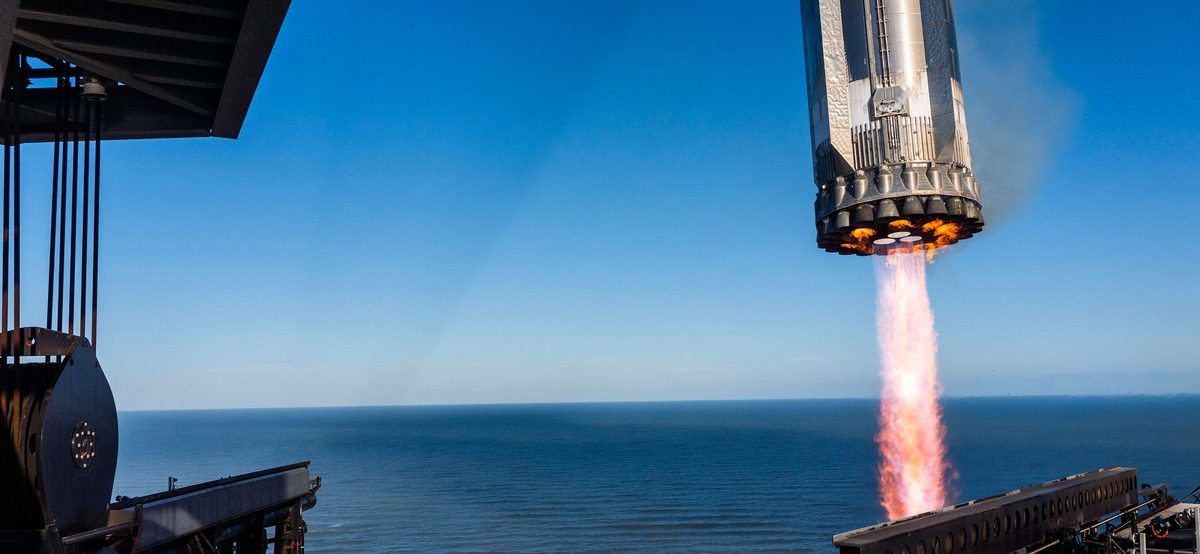 Seen from the top of the launch tower, the Super Heavy booster comes down tail-first with its engines firing, parts of the tower in the foreground and the sea behind