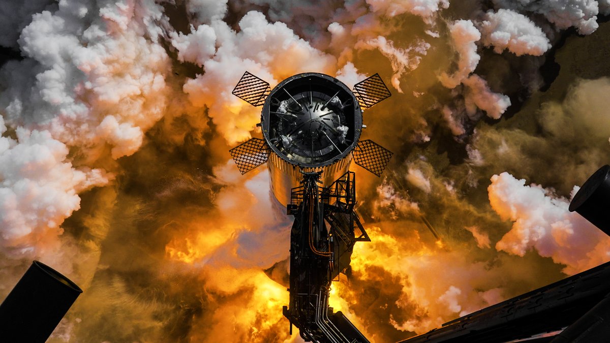 Looking down on the top of a Super Heavy booster, its four lattice fins spread, as flame and white clouds from its engines billow around the launch mount