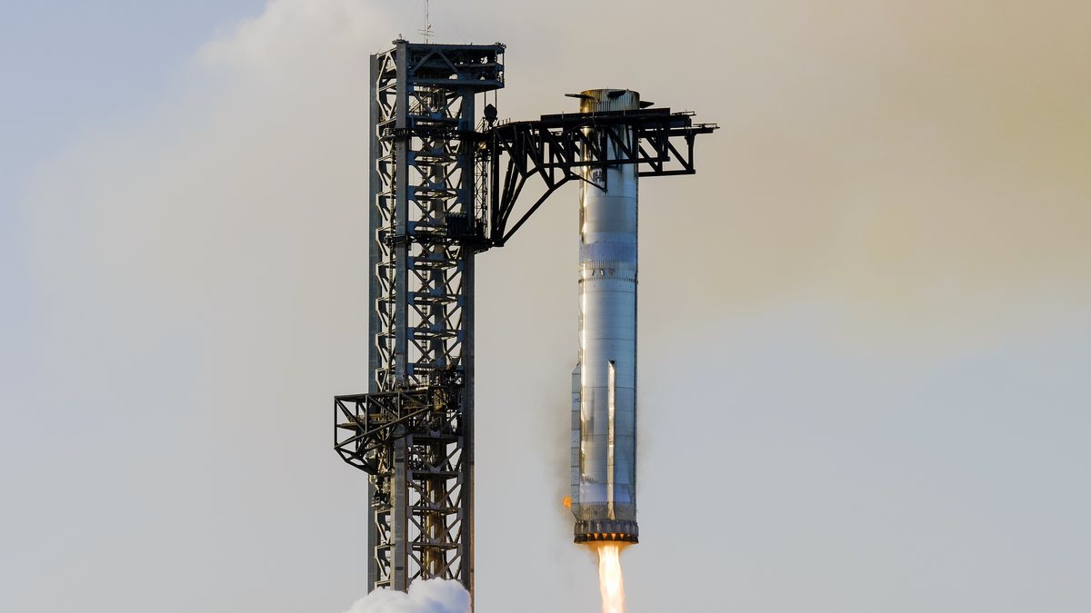 The Super Heavy booster, engines still firing, held at its top by the two arms of the launch tower