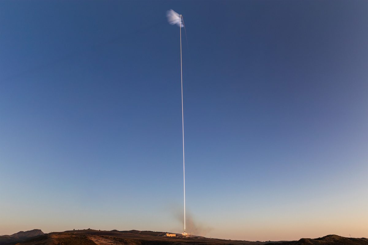 A thin bright line rising almost straight up from a hilltop launch pad into a clear blue evening sky, with a small white cloud at the top of the line and an orange glow on the horizon