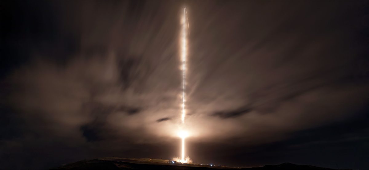 A bright vertical streak of light rising from a glowing launch site on a dark hill into a night sky with long streaks of cloud